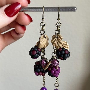 May include: Handmade earrings with a blackberry theme. The earrings feature clusters of dark purple and black berries, tan leaves, and dangling purple beads. The earrings are suspended from a silver bar.