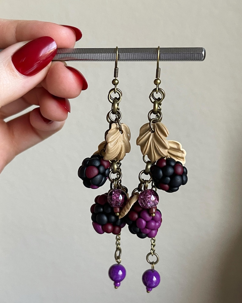 May include: Handmade earrings with a blackberry theme. The earrings feature clusters of dark purple and black berries, tan leaves, and dangling purple beads. The earrings are suspended from a silver bar.