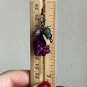 May include: A handmade earring featuring a cluster of purple beads resembling grapes, accented with green and pink beads. The earring is suspended from a gold-coloured hook and chain. The earring is approximately 3.8 cm long.