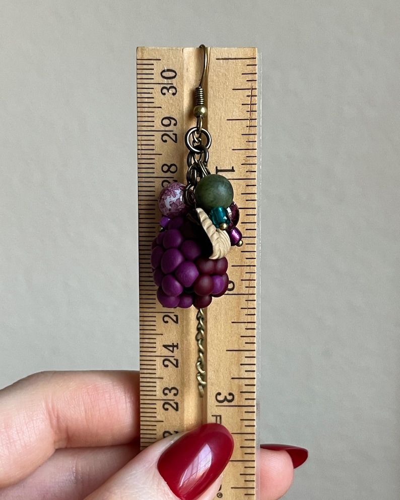 May include: A handmade earring featuring a cluster of purple beads resembling grapes, accented with green and pink beads. The earring is suspended from a gold-coloured hook and chain. The earring is approximately 3.8 cm long.