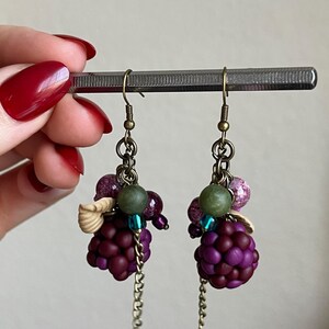 May include: Handmade earrings with dark purple grape-like bead clusters, complemented by green, purple, and blue beads, small seashell details, and dangling chains. The ear wires are antique brass.
