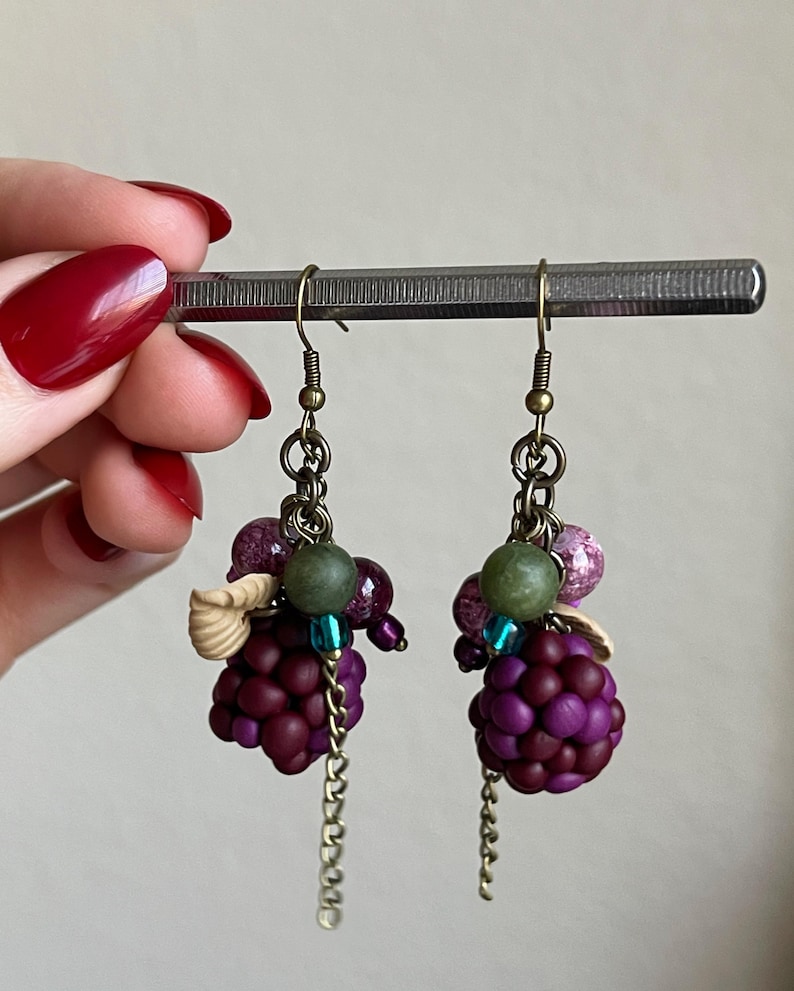 May include: Handmade earrings with dark purple grape-like bead clusters, complemented by green, purple, and blue beads, small seashell details, and dangling chains. The ear wires are antique brass.