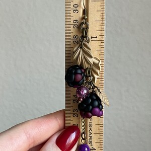 May include: A handcrafted earring with a blackberry design, featuring black and purple charms, pink beads, and tan leaf accents. The earring is about 7.6 cm long, with a bronze-toned hook and chain.