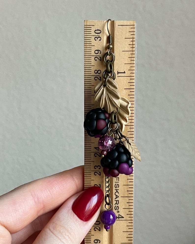 May include: A handcrafted earring with a blackberry design, featuring black and purple charms, pink beads, and tan leaf accents. The earring is about 7.6 cm long, with a bronze-toned hook and chain.
