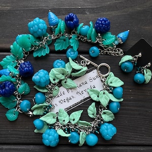 May include: A handmade bracelet and matching earrings featuring blue and turquoise berry-shaped beads and green leaf accents. The jewelry is connected by silver-colored chains. The text on the tag reads "made by Lana Sun, Las Vegas, Nevada."