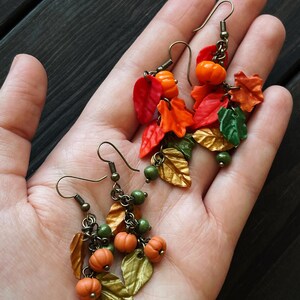 May include: A pair of dangle earrings with a fall theme. The earrings feature small orange pumpkins, green beads, and gold and orange leaves.