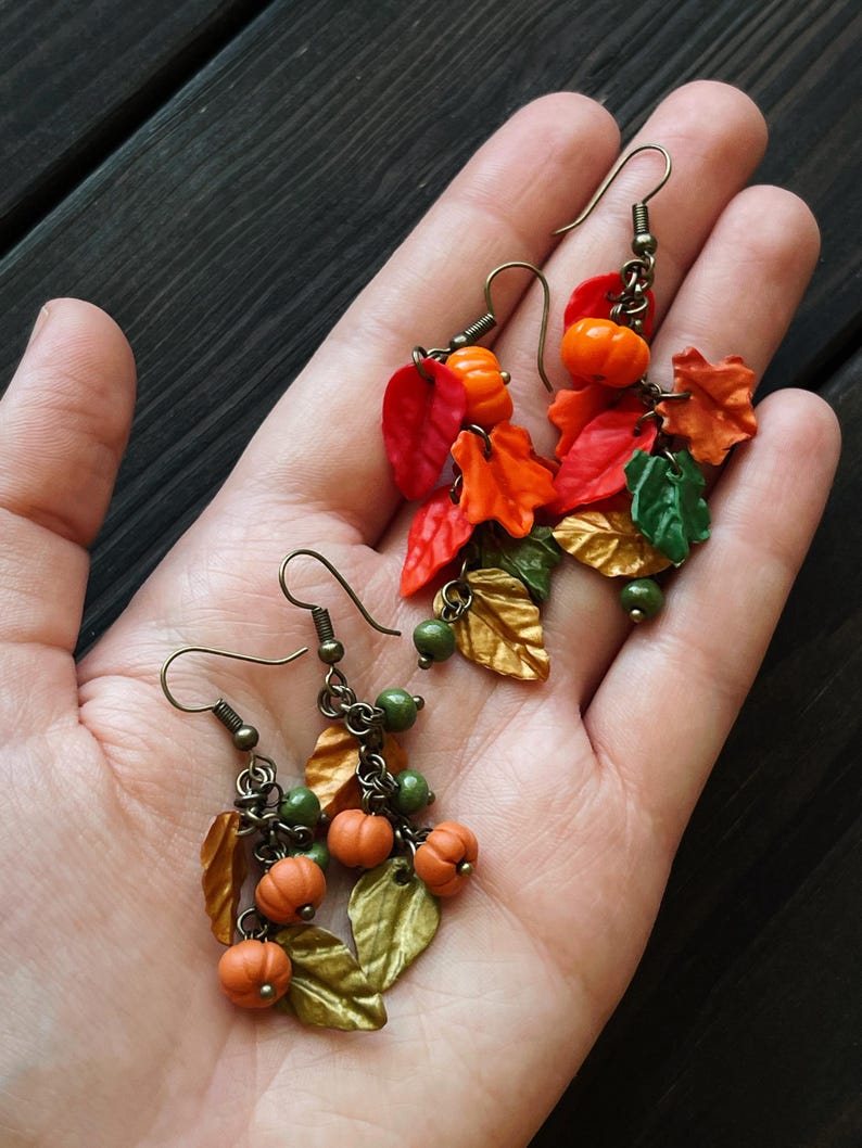 May include: A pair of dangle earrings with a fall theme. The earrings feature small orange pumpkins, green beads, and gold and orange leaves.