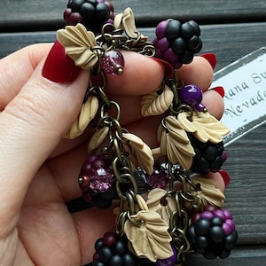 May include: Handmade earrings with blackberry clusters in purple and black, tan leaves, and purple beads. The earrings hang from a bronze chain. The design evokes a natural, rustic aesthetic.