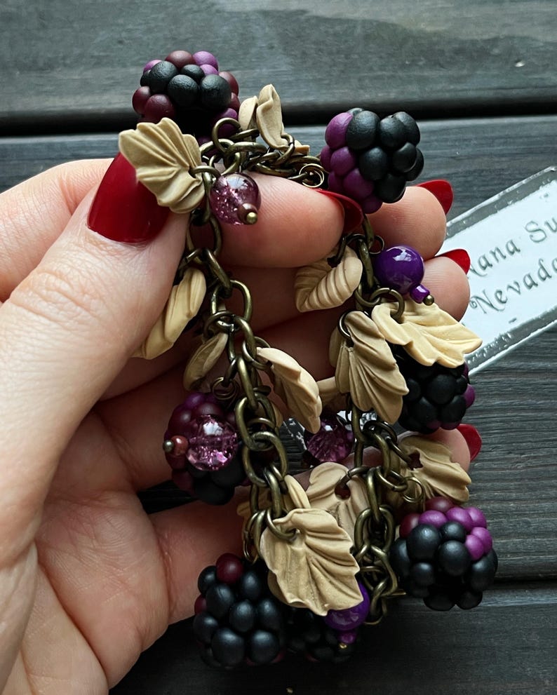 May include: Handmade earrings with blackberry clusters in purple and black, tan leaves, and purple beads. The earrings hang from a bronze chain. The design evokes a natural, rustic aesthetic.