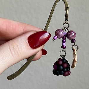 May include: A bronze metal bookmark with a decorative charm. The charm includes purple beads, a cluster of black and red beads, and a small tan paper element. The bookmark is held by a hand with red nail polish.