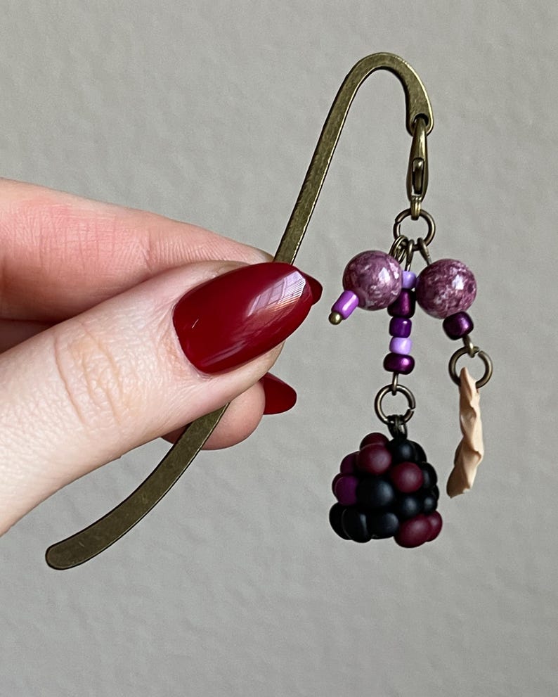 May include: A bronze metal bookmark with a decorative charm. The charm includes purple beads, a cluster of black and red beads, and a small tan paper element. The bookmark is held by a hand with red nail polish.