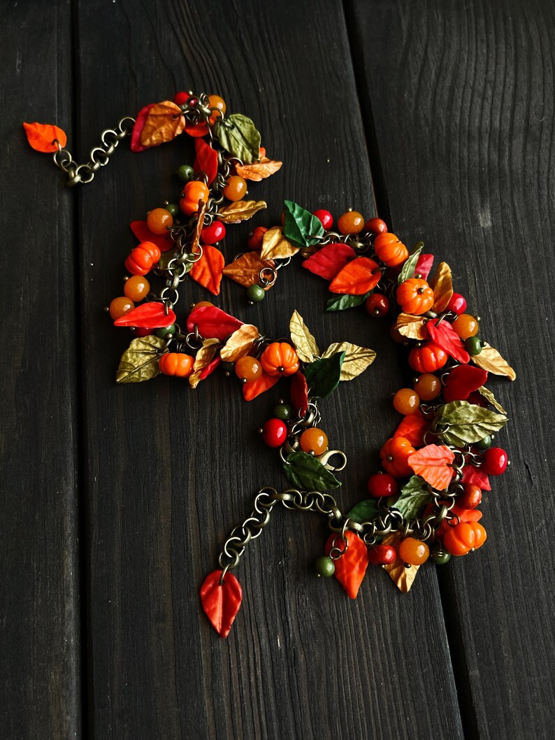 May include: A fall-themed necklace with orange, red, and gold leaves, small orange pumpkins, and brown beads. The necklace is made of metal chain and polymer clay.