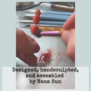May include: A craft project in progress is shown. Three small, red, berry-like objects are impaled on a needle. A pink brush is used to apply a red powder. Text reads: Designed, handsculpted, and assembled by Nana Sun.