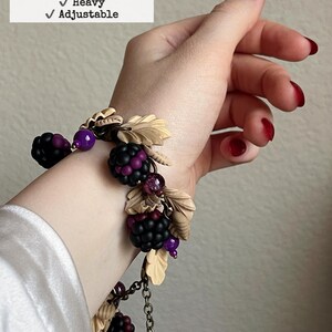 May include: A hand-sculpted bracelet with brass and copper alloy elements, polyclay, glass, and acrylic. It features black and purple berry clusters, beige leaves, and purple beads. The bracelet is 21.6 cm long, heavy, and adjustable.