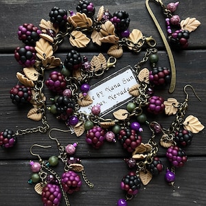 May include: A jewellery set with a blackberry theme, including a bracelet, earrings, and a bookmark. The pieces feature dark purple, black, and green beads, and tan leaf accents. The jewellery is displayed on a dark wooden surface. The set is handmade.