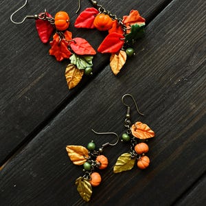 May include: A pair of dangle earrings with a fall theme. The earrings feature small orange pumpkins, gold and green leaves, and brown chain links.