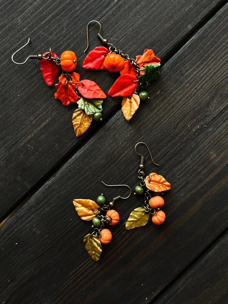 May include: A pair of dangle earrings with a fall theme. The earrings feature small orange pumpkins, gold and green leaves, and brown chain links.