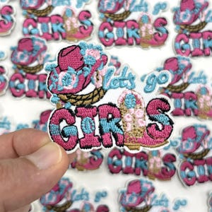 May include: Embroidered patch with the text "Let's go GIRLS" in pink and blue. The patch features a pink cowboy hat with blue floral accents and a brown woven basket. The patches are on a white background.