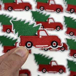 May include: Red truck patch with a green Christmas tree in the truck bed. The patch is embroidered with white and black details. Many similar patches are in the background.