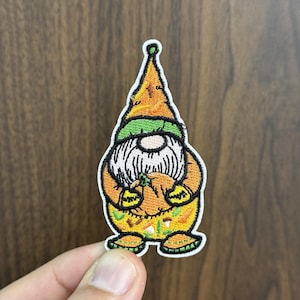 May include: Embroidered patch featuring a gnome with a long white beard, wearing an orange hat and holding a pumpkin. The gnome has a green hat band and a yellow and green patterned outfit. The patch is on a wooden surface.