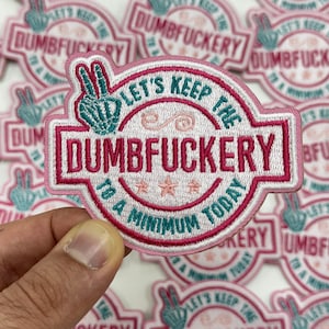 May include: Pink and white embroidered patch with the text "LET'S KEEP THE DUMBFUCKERY TO A MINIMUM TODAY". The patch features a peace sign and decorative elements in teal and pink.