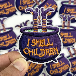 May include: Embroidered patch featuring a purple cauldron with the text "I SMELL CHILDREN" in orange. The cauldron has green liquid and witch legs sticking out. A hand holds the patch, surrounded by similar patches.