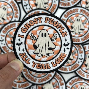May include: Round patch with the text "I Ghost People All Year Long" and a cartoon ghost giving a peace sign. The patch has a white border, orange and white checkered background, and black accents.