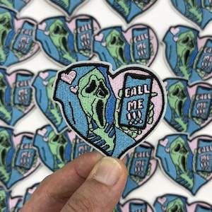 May include: Embroidered patch featuring a Scream mask holding a phone with "CALL ME" on the screen, set within a blue heart with pink accents. The patch is held by a hand, with several similar patches in the background.