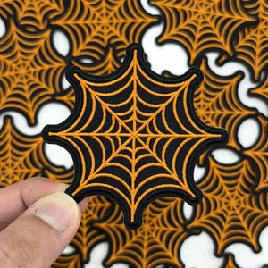 May include: Black and orange spiderweb patch with an embroidered design. The patch has a black background with an orange spiderweb design. The patch is held in a hand, with other patches in the background.