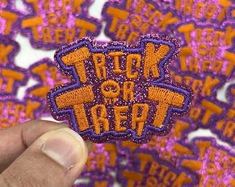 Può includere: Primo piano di una toppa a tema Halloween con le parole "TRICK OR TREAT" in arancione, bordate di viola e impreziosite da glitter. Una piccola grafica a teschio è tra le parole. Lo sfondo è pieno di toppe simili.