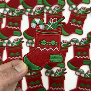 May include: Red and green Christmas stocking patch with white zigzag stitching and embroidered details. The stocking is filled with a candy cane and a wrapped gift. The background features multiple similar patches, creating a festive holiday theme.
