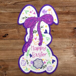 May include: A white felt Easter bunny with a purple sequin bow and the text "Happy Easter" on a wooden background.