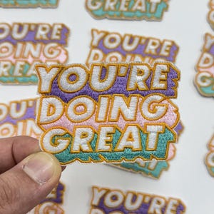 May include: Embroidered patches with the phrase "YOU'RE DOING GREAT" in white, outlined in gold, and set against a purple, pink, and teal background. The patches are displayed on a light background.