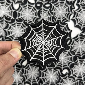 May include: A black and white spiderweb patch with a detailed white web design. The patch is held against a background of spiderweb patterns. This patch is perfect for Halloween or spooky-themed crafts.