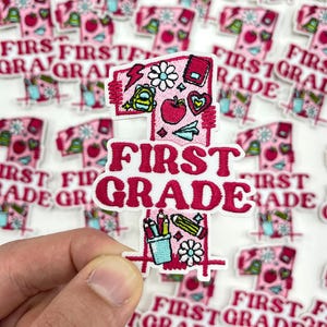 May include: Pink and white embroidered patch with the text "FIRST GRADE". The patch features a number one design with school-themed illustrations like an apple, backpack, and pencils. The background is covered with similar patches.