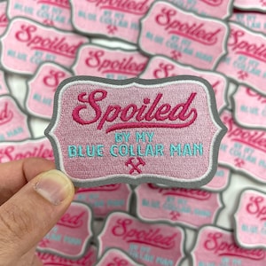 May include: A close-up of a pink and gray embroidered patch with the text "Spoiled by my Blue Collar Man" in pink and blue lettering. The patch has a gray border and a small crossed hammer and pickaxe design in the bottom center.