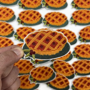 May include: Embroidered patch featuring a detailed pie design with a lattice crust and cherry filling. The patch has a green border and is surrounded by other similar patches, creating a repeating pattern. The pie patches are approximately 2 inches in diameter.