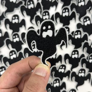 May include: A black ghost patch with a white border is held in front of a background of similar ghost patches. The patch has a simple design with a white face and a flowing body, perfect for Halloween.