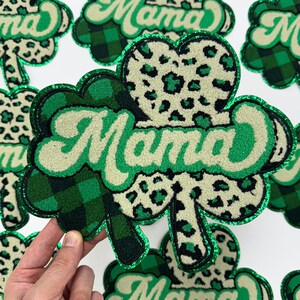 May include: Shamrock-shaped patches with the word "Mama" in green script. The patches feature a mix of green plaid and leopard print patterns. The edges are outlined with a glittery green trim.