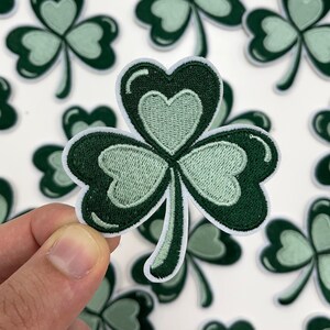 May include: A close-up of a green shamrock patch with a white outline. The shamrock is made of felt and has a three-dimensional design.
