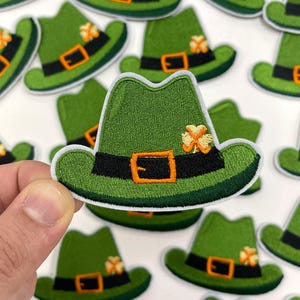 Puede incluir: Una colección de sombreros irlandeses de duende en fieltro verde con ribetes naranja y negro y un diseño de trébol. Los sombreros están bordados con un cinturón negro y una hebilla.