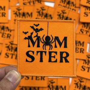 May include: Orange square patch with black text and Halloween-themed graphics. The patch reads "MOM STER" with a spider hanging from a string and bats above. The patch is surrounded by other similar patches.