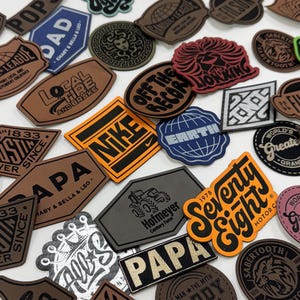 May include: A collection of various leather patches in different shapes and colors. Some patches feature text like "NIKE," "PAPA," and "Seventy Eight." Other patches have graphic designs, including a lion and a globe.