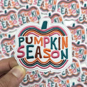 May include: Embroidered patch in the shape of a pumpkin with the words "PUMPKIN SEASON" in teal, orange, and pink. The patch has a white border and a green stem. The background shows multiple similar patches.