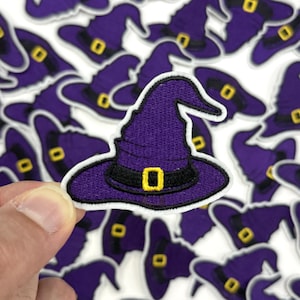 May include: A collection of purple witch hat patches with black bands and gold buckles. The patches are embroidered with a white border. One patch is held up by a hand, with many more patches in the background.
