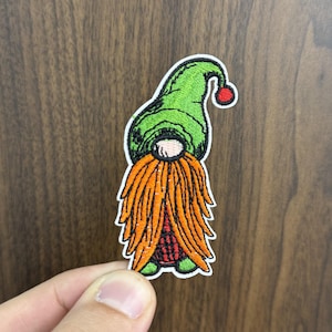 May include: Embroidered patch featuring a whimsical gnome design. The gnome has a green hat with a red pom-pom, a long orange beard, and a white face. The patch has a white border and is likely for clothing or accessories.