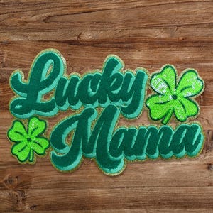 May include: Green felt patch with the words "Lucky Mama" in a cursive font with gold glitter accents. Two green shamrock patches are on either side of the text.