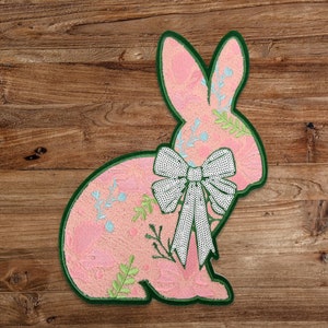 May include: A pink felt bunny with a green outline and a white bow with sequins. The bunny is decorated with a floral pattern in pink, green, and blue.