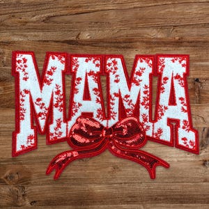 May include: White felt patch with the word "MAMA" in red outlined letters. The letters are covered in a small floral pattern. A red sequined bow is attached to the bottom of the patch.