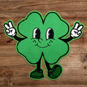 May include: A green four-leaf clover with a smiling face and two arms raised in a peace sign. The clover has a cartoon-like appearance and is made of a soft, plush material.